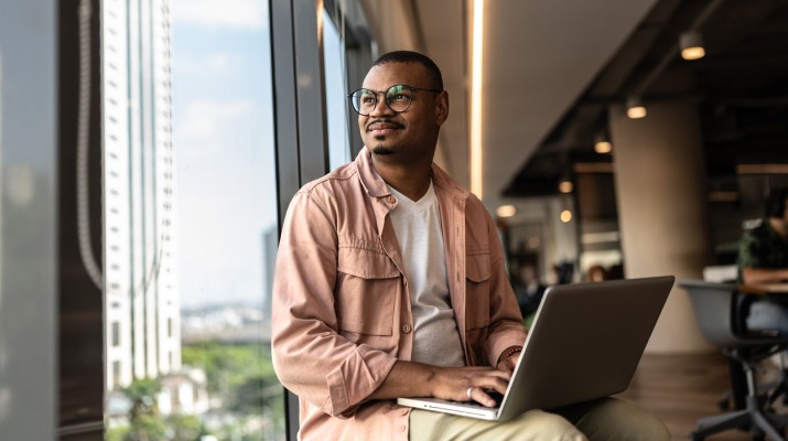 A man sitting with a laptop smiling and looking out of the window.
